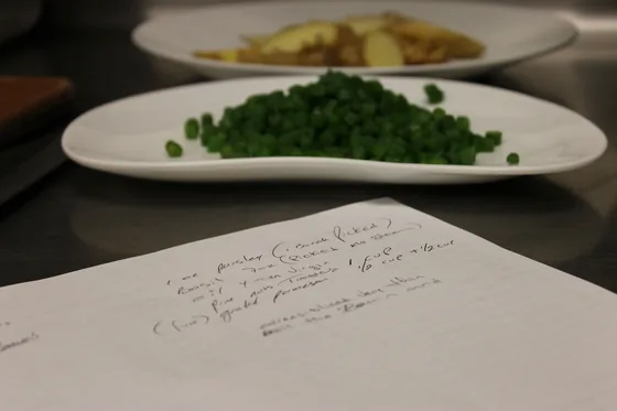 A handwritten recipe sheet on the pass beside plates of peas and potatoes.