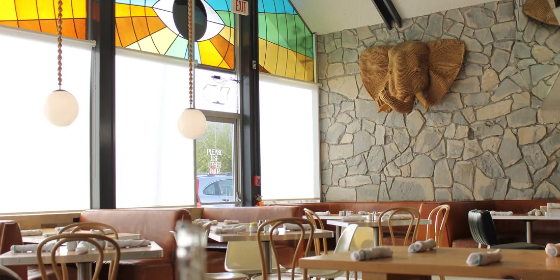 An empty dining room in daylight: stained glass above tall windows, a stone wall, set tables.